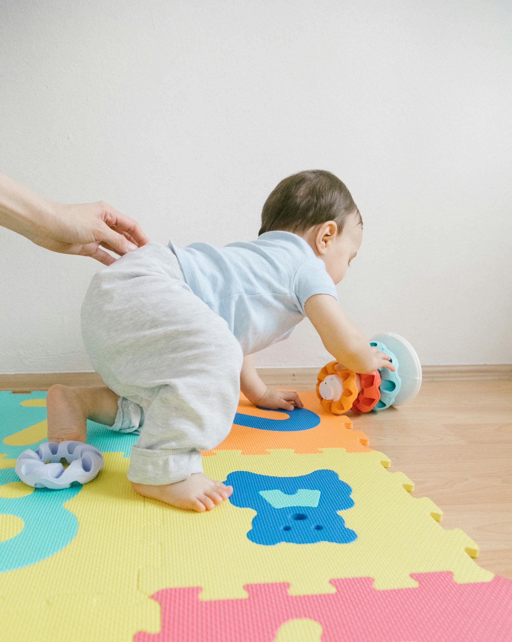 A toddler playing with colorful toys on a puzzle mat indoors, guided by an adult hand.
