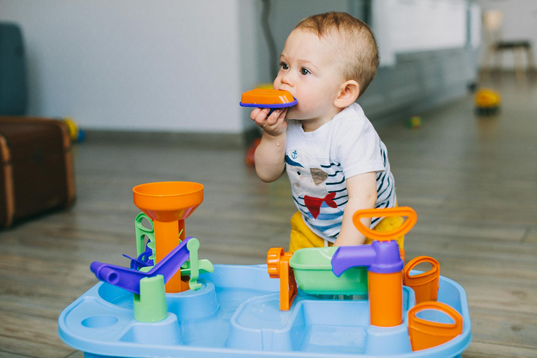 Adorable toddler chewing on toy piece while playing indoors with bright colored plastic toy.