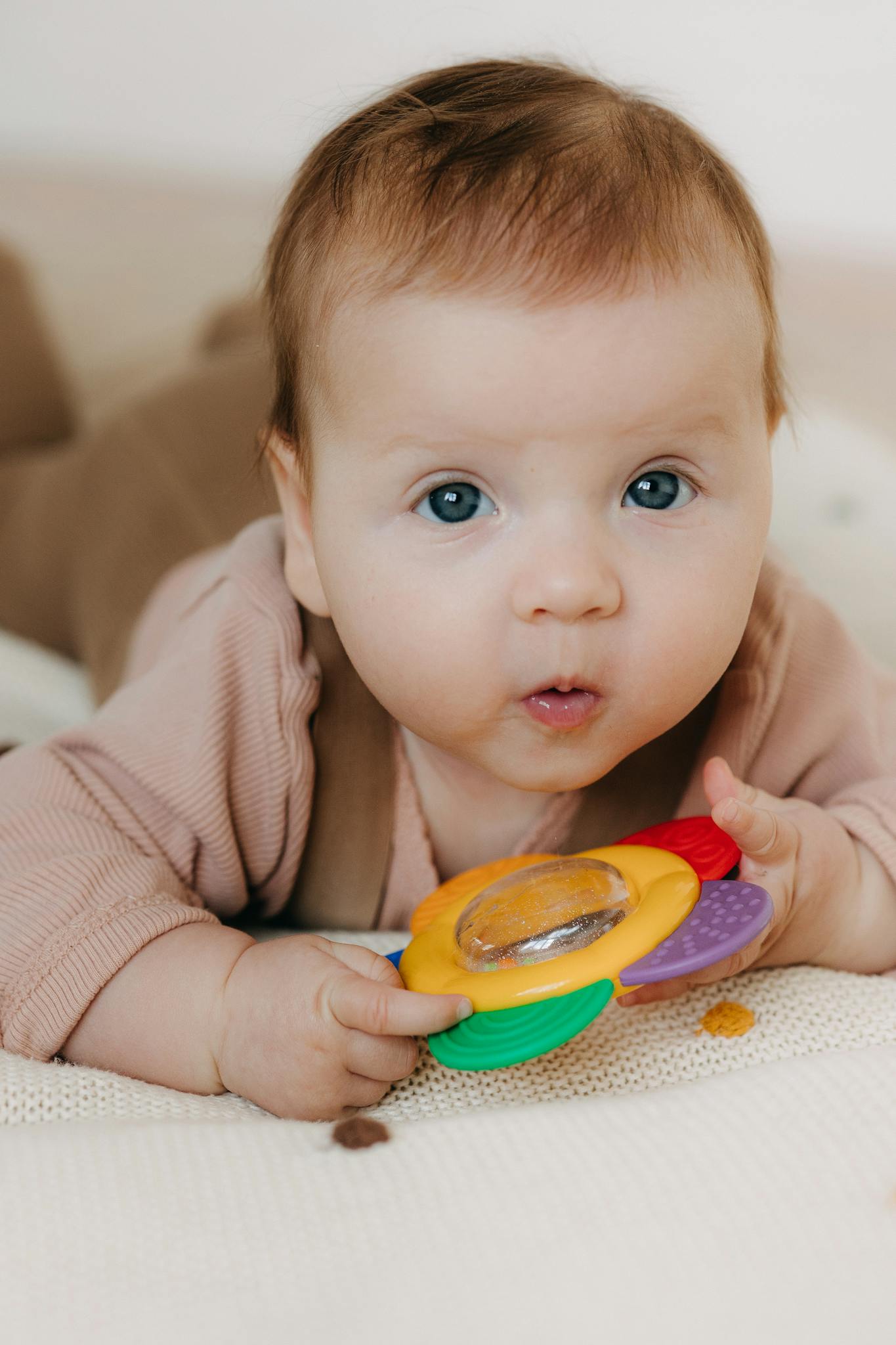 Cute baby holding a colorful sensory toy while lying on a soft surface indoors.